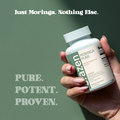 Hand holding a bottle of Brazen Moringa Pure capsules with text on a green background